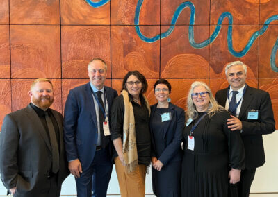 A group of people posing together in front of an art piece (a multi-tiled sculpted painting with a dark brown background, lighter brown hills, and a blue river) at Parliament House. This group includes Nick Verginis, the CEO of SENVIC.