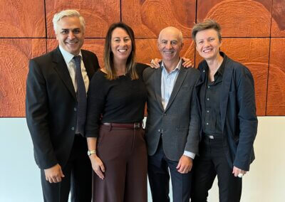 A group of people posing together in front of an art piece (a multi-tiled sculpted painting with a dark brown background, lighter brown hills, and a blue river) at Parliament House. This group includes Nick Verginis, the CEO of SENVIC.