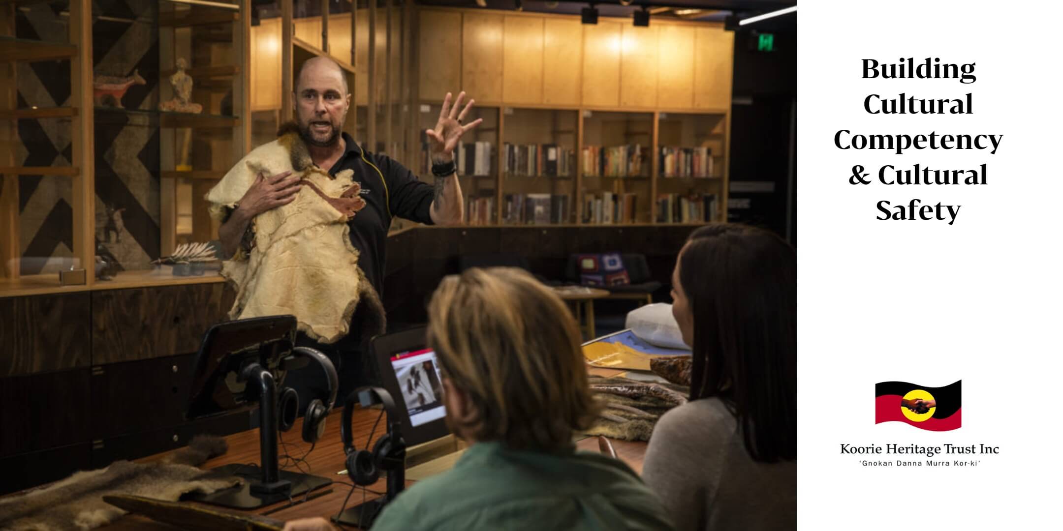 In a photo on the left of this banner a man holding a possum skin is talking to a class of people in a dimly lit library or museum. He is holding his hand up as though he is gesticulating, explaining or emphasising a point. On the right are the words “Building Cultural Competency and Cultural Safety” and the logo for Koorie Heritage Trust inc - ‘Gnomon Danna Murray Kor-ki’